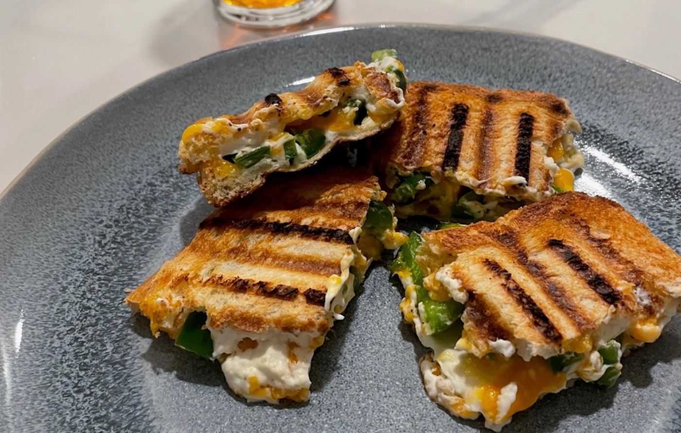 Grilled jalapeño popper sandwich cut in half, cheese melting out, served with a glass of beer for game day.