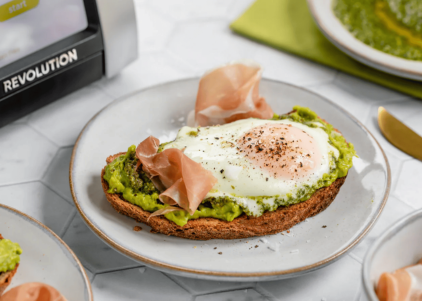 Golden artisan toast topped with creamy avocado, a pesto-fried egg, and thin slices of prosciutto, garnished with red pepper flakes.