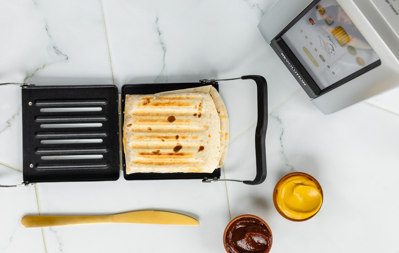 Revolution Toastie Press open on the counter with a toasted pocket fold quesadilla inside. a knife, bbq sauce, and mustard next to it, the toaster is above it
