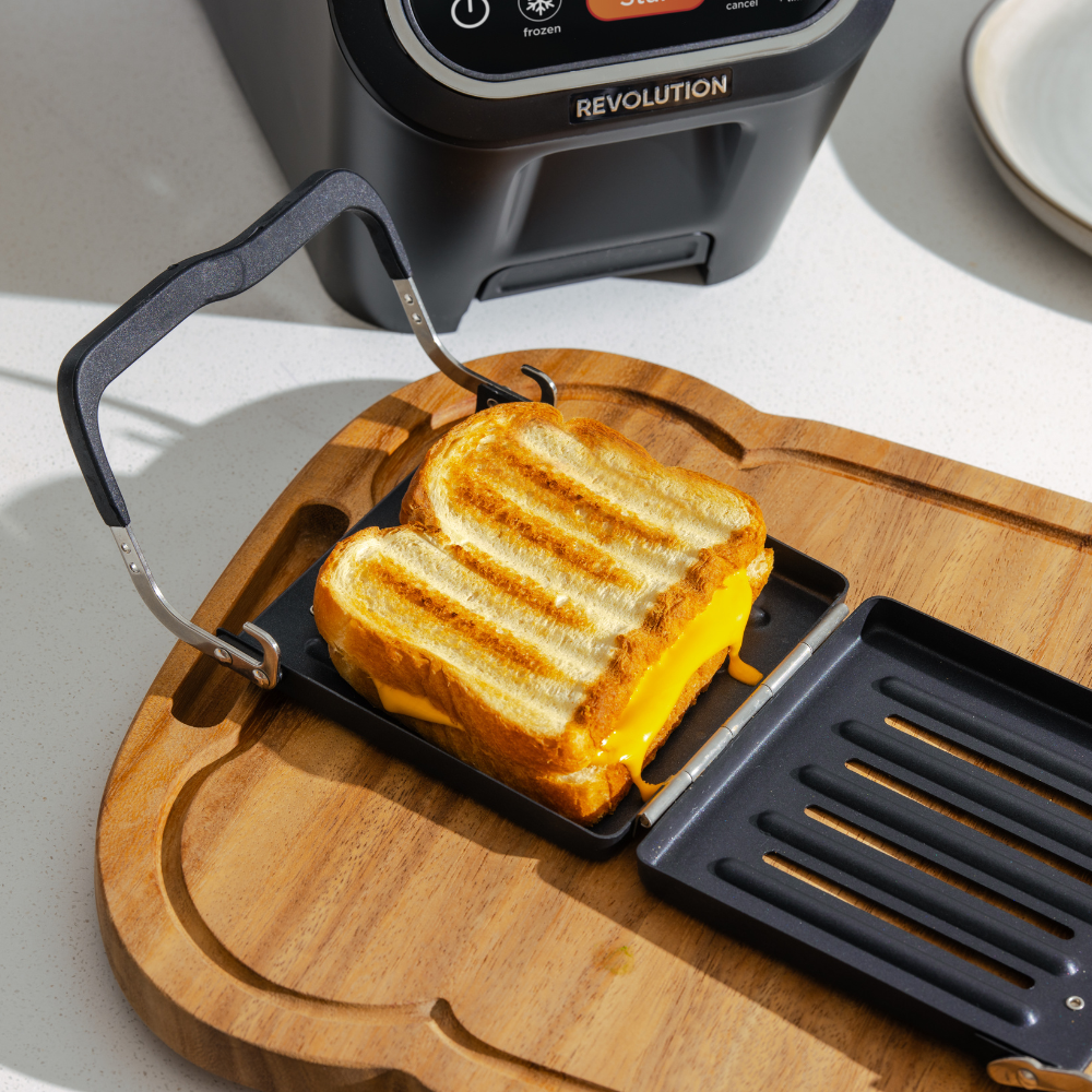 A grilled cheese sandwich with melted cheddar is being removed from the Revolution Cooking R90 Digital Toaster, featuring a digital screen, onto a wooden cutting board. The precise temperature control ensures perfection every time, with a plate conveniently placed nearby for serving.