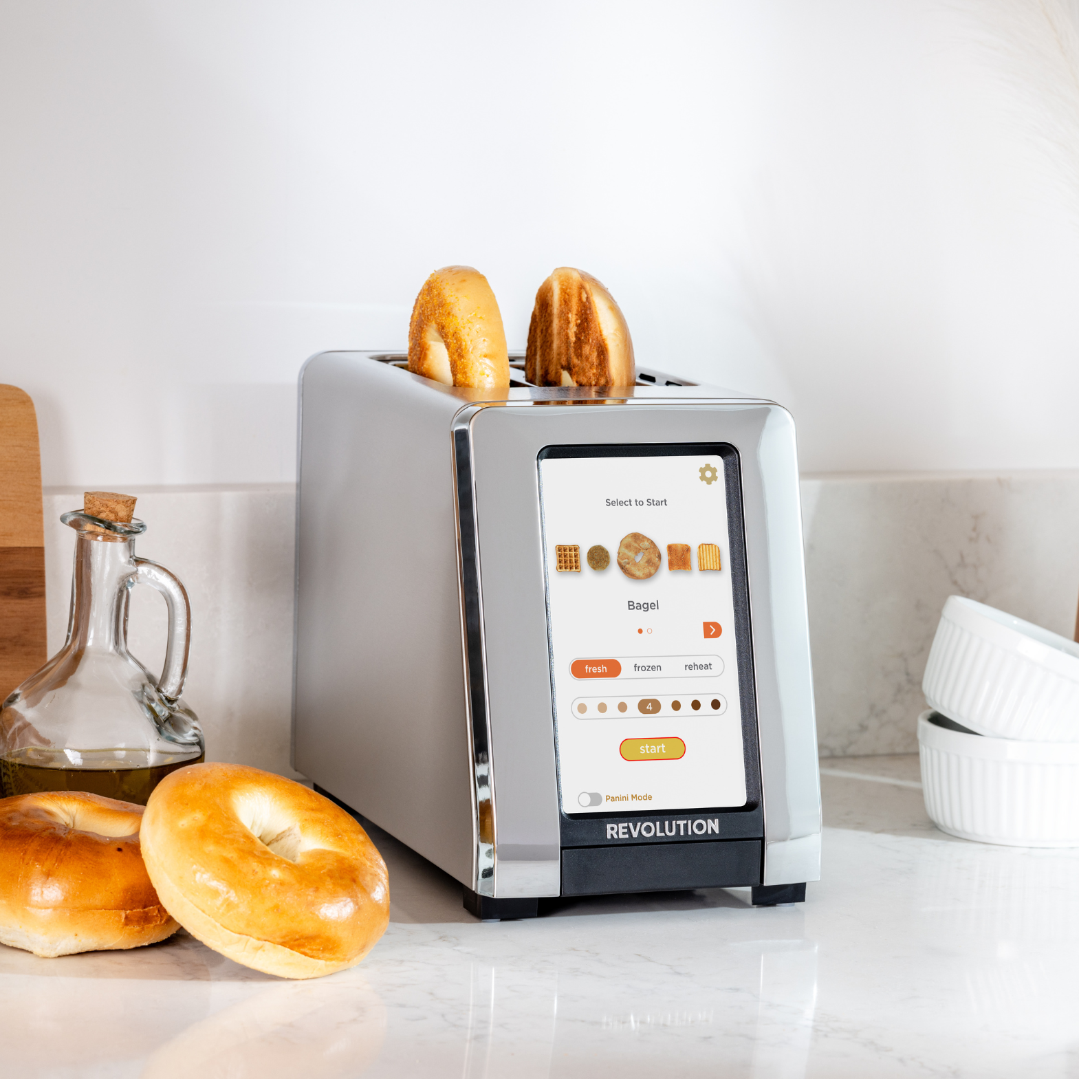 The Renewed R180 Smart Toaster by Revolution Cooking showcases its InstaGLO heating system with bagels being toasted on a digital display, offering options like Panini Mode. Two bagels are inside and one nearby, while olive oil and stacked dishes adorn the counter.