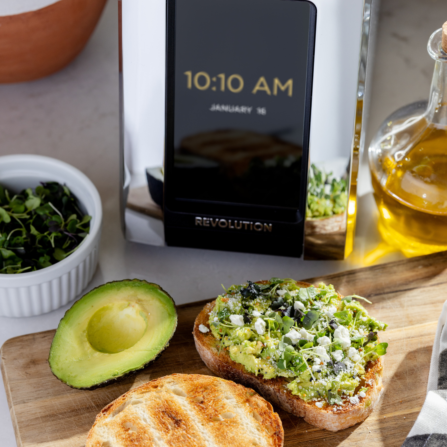 A toasted slice of bread with avocado and greens sits on a wooden board next to half an avocado and a bowl of microgreens. The Renewed R180 Smart Toaster by Revolution Cooking shows 10:10 AM, January 16, while a container of olive oil is nearby.