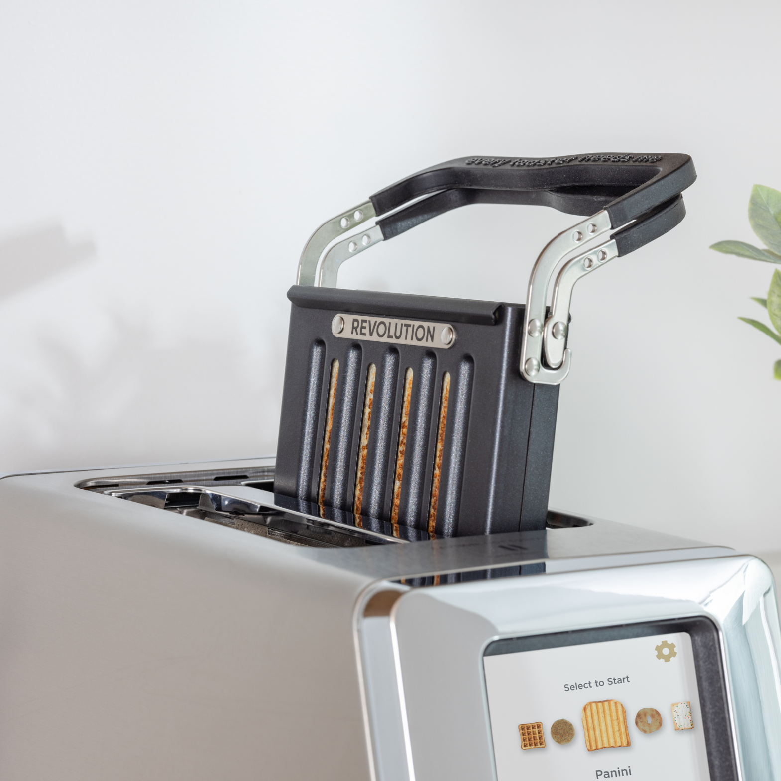 The Renewed R180 Smart Toaster by Revolution Cooking features a touchscreen and the cutting-edge InstaGLO heating system. It includes a "Revolution" metallic insert and seems ideal for making sandwiches. A small plant can be seen in the background.