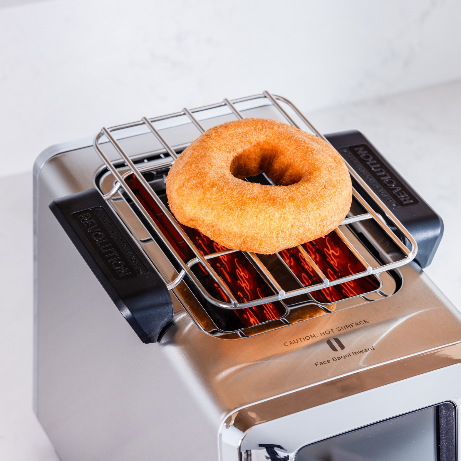 A single bagel rests on a metal warming rack atop a Renewed R180 Smart Toaster by Revolution Cooking, featuring the InstaGLO heating system and a digital display. The toaster is placed on a white countertop, with the rack labeled for warming bagels.