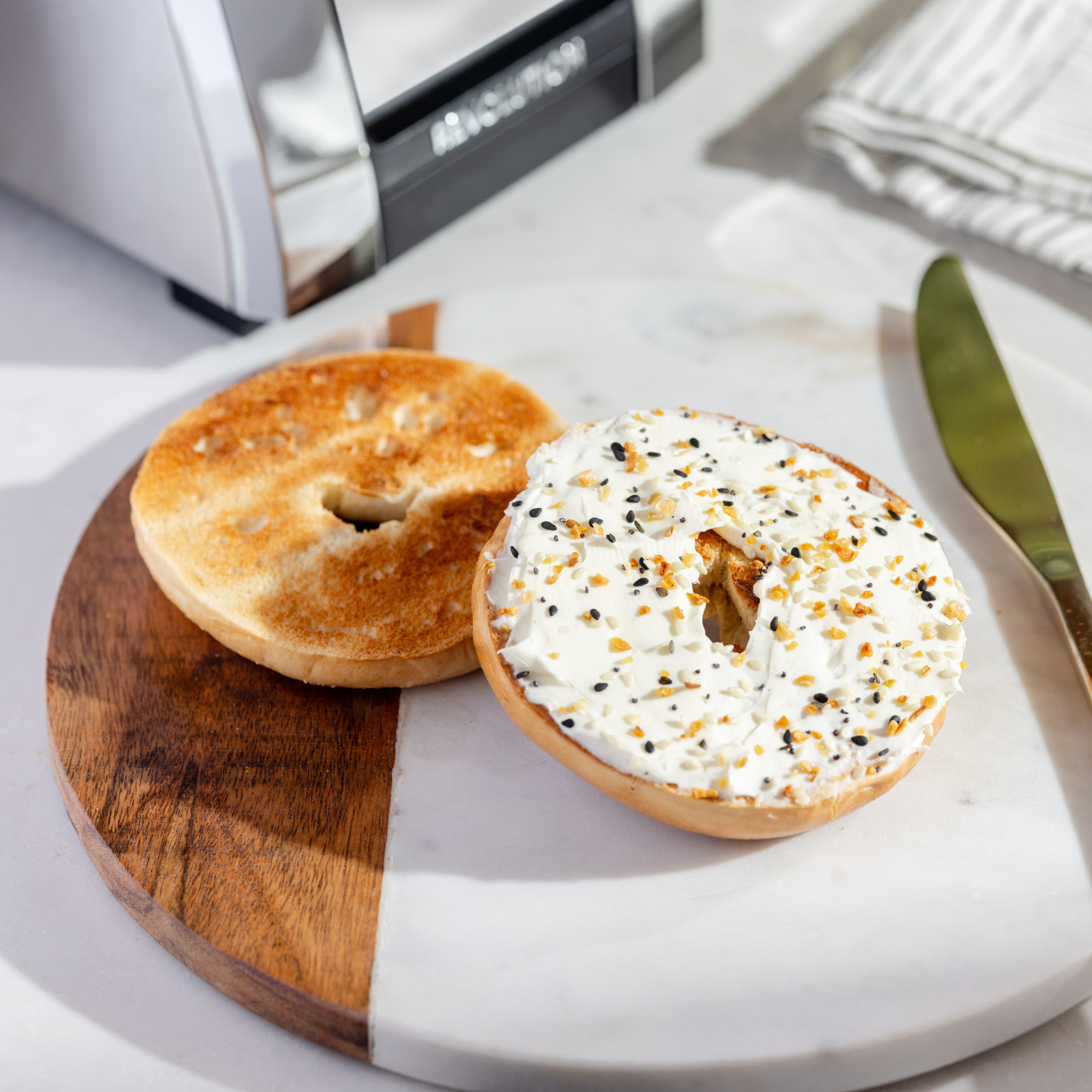 A wooden and marble board holds a halved bagel: one plain, one with cream cheese and everything seasoning, freshly made using the Renewed R180 Smart Toaster by Revolution Cooking. A knife rests nearby, with this sophisticated toaster featured in the background.