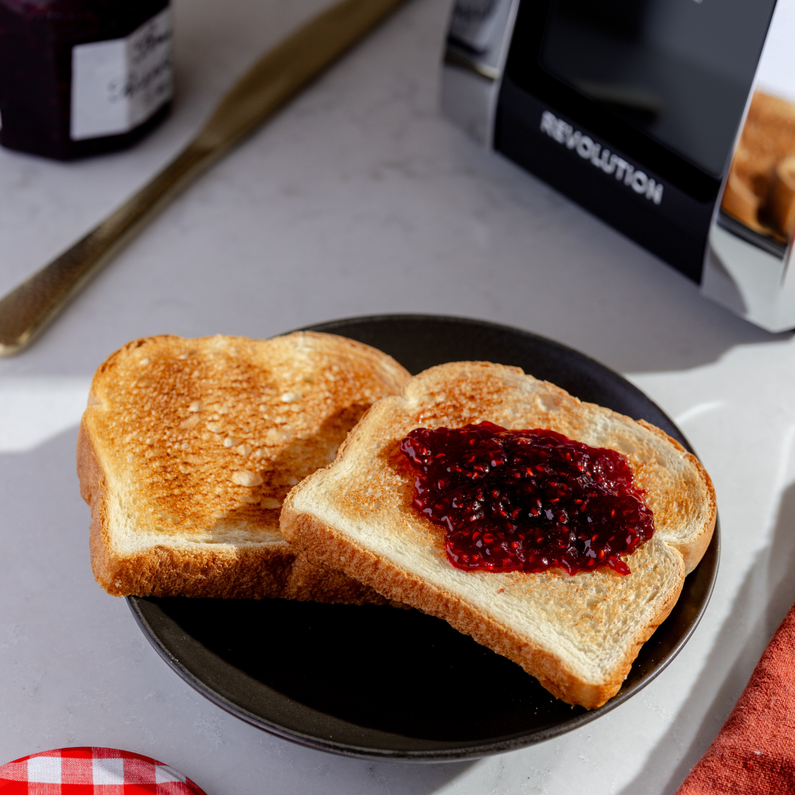 Two slices of toast rest on a dark plate, one with red jam. Nearby are a jam jar and a Renewed R180 Smart Toaster by Revolution Cooking featuring the InstaGLO heating system. A red-and-white checkered cloth adds a cozy touch to the scene.