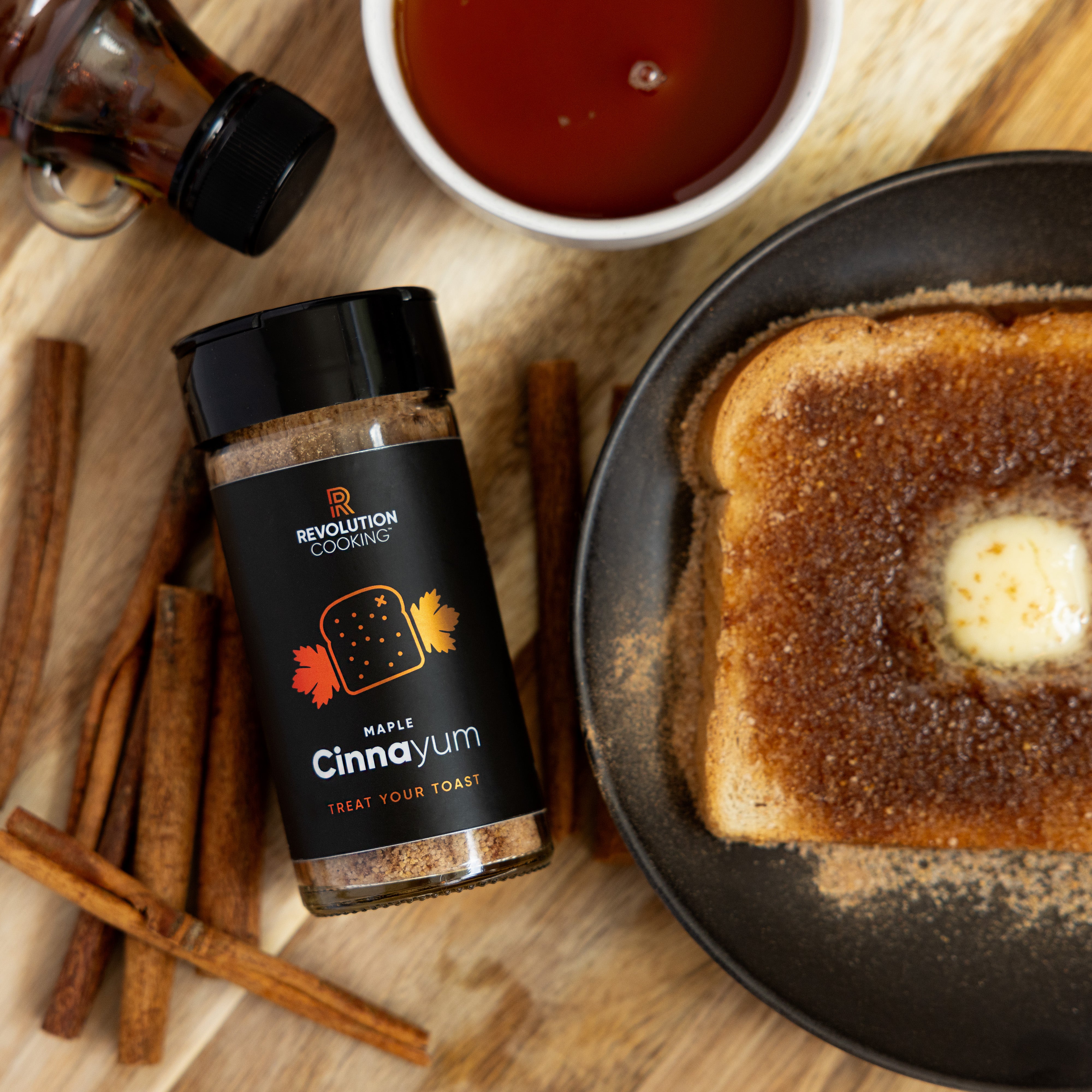 A jar of Maple Cinnayum from Ocean State Pepper Co. sits beside a slice of toast covered in melted butter and cinnamon sugar. Nearby, cinnamon sticks and a cup of brown liquid rest on the wooden surface, creating the perfect vanilla-infused morning indulgence.