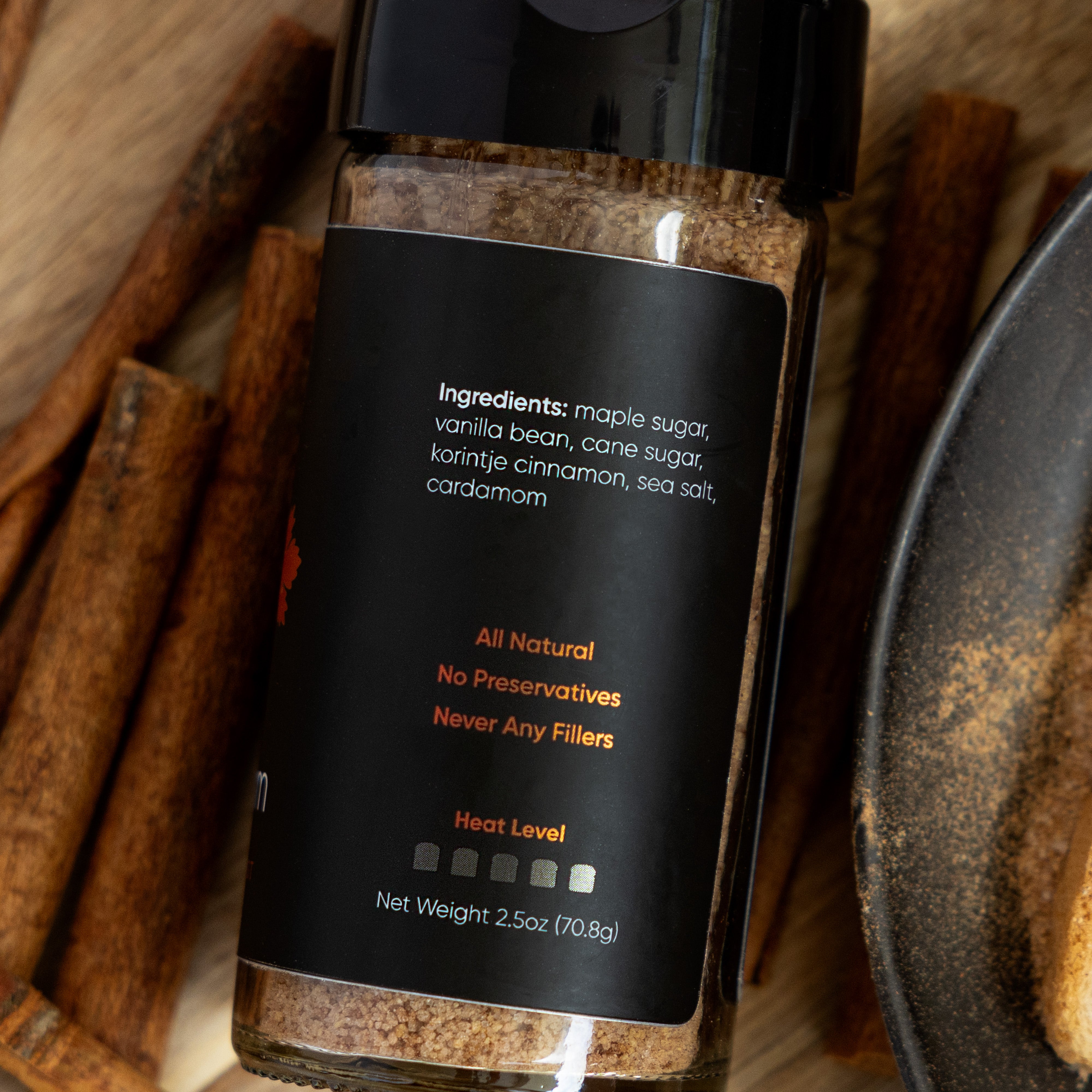 Displayed is a jar of seasoning by Ocean State Pepper Co., named Maple Cinnayum. The jar features a black label with bold statements: "All Natural," "No Preservatives," and "Never Any Fillers." The ingredients listed are cinnamon maple sugar, vanilla bean, cane sugar, and spices. Cinnamon sticks nearby enhance the rich, vanilla-infused aroma.