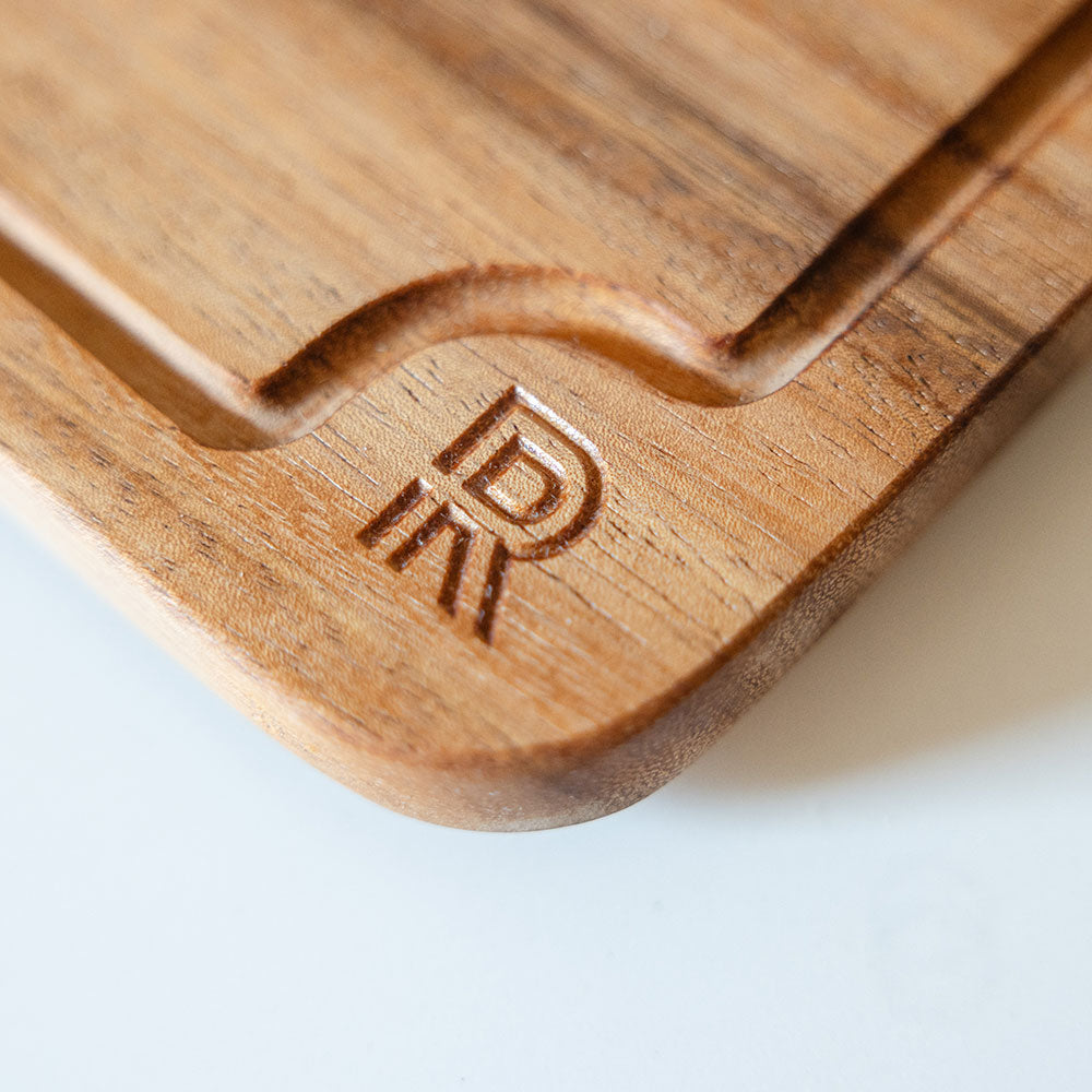 Close-up of the corner of the Hardenbrook Hardwoods cutting board, meticulously crafted from luxurious monkey wood. It features a smoothly carved groove and is engraved with the initials "DR". The wood grain is prominently visible, showcasing the board's exceptional craftsmanship.