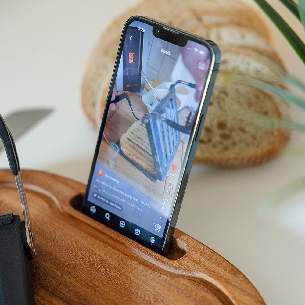 A smartphone, nestled in a Hardenbrook Hardwoods Cutting Board with a phone slot, displays a video of a sandwich being grilled. In the background, a loaf of bread rests on the monkey wood surface.