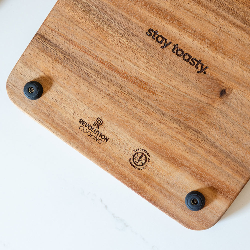 A cutting board crafted from smooth, light-colored monkey wood and engraved with the phrase "stay toasty." It features the brand "Hardenbrook Hardwoods," includes non-slip feet and a convenient phone slot, and is elegantly presented on a white background.