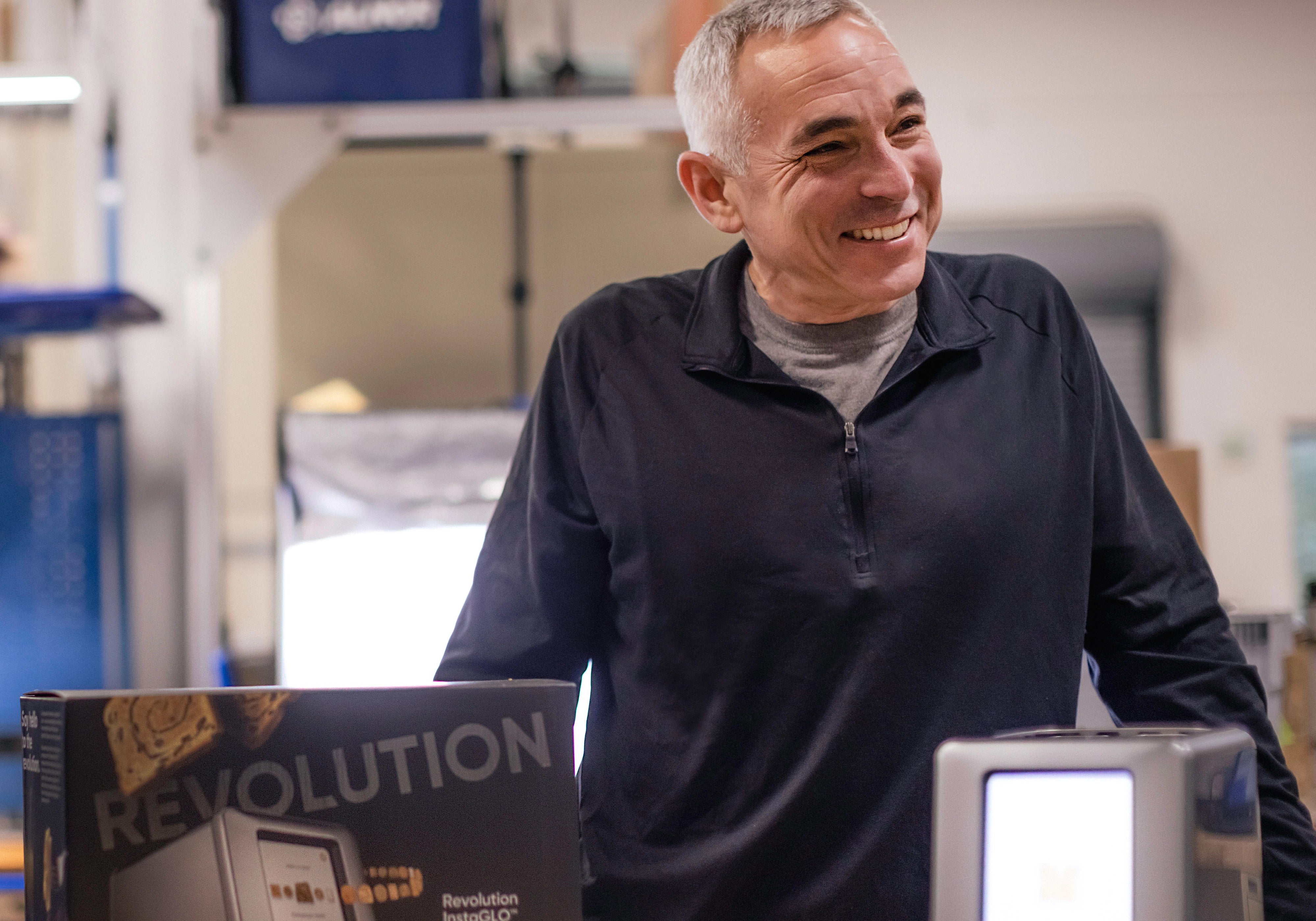 A person with short gray hair is smiling while standing indoors near a product box labeled "Revolution." The environment appears to be an industrial or workshop setting.