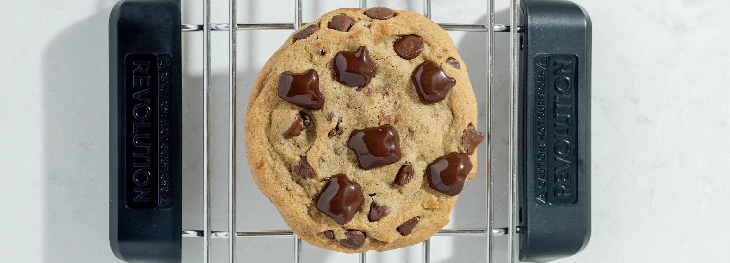 A chocolate chip cookie with melted chocolate chunks on a cooling rack, viewed from above. The rack rests on a light surface, and the cookie is centered in the image.