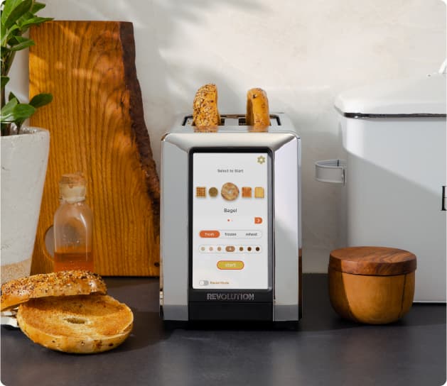 A toaster with digital controls toasts two bagel halves. A bagel with sesame seeds is placed nearby on a wooden cutting board. A small plant, honey jar, and wooden containers are arranged on the counter.