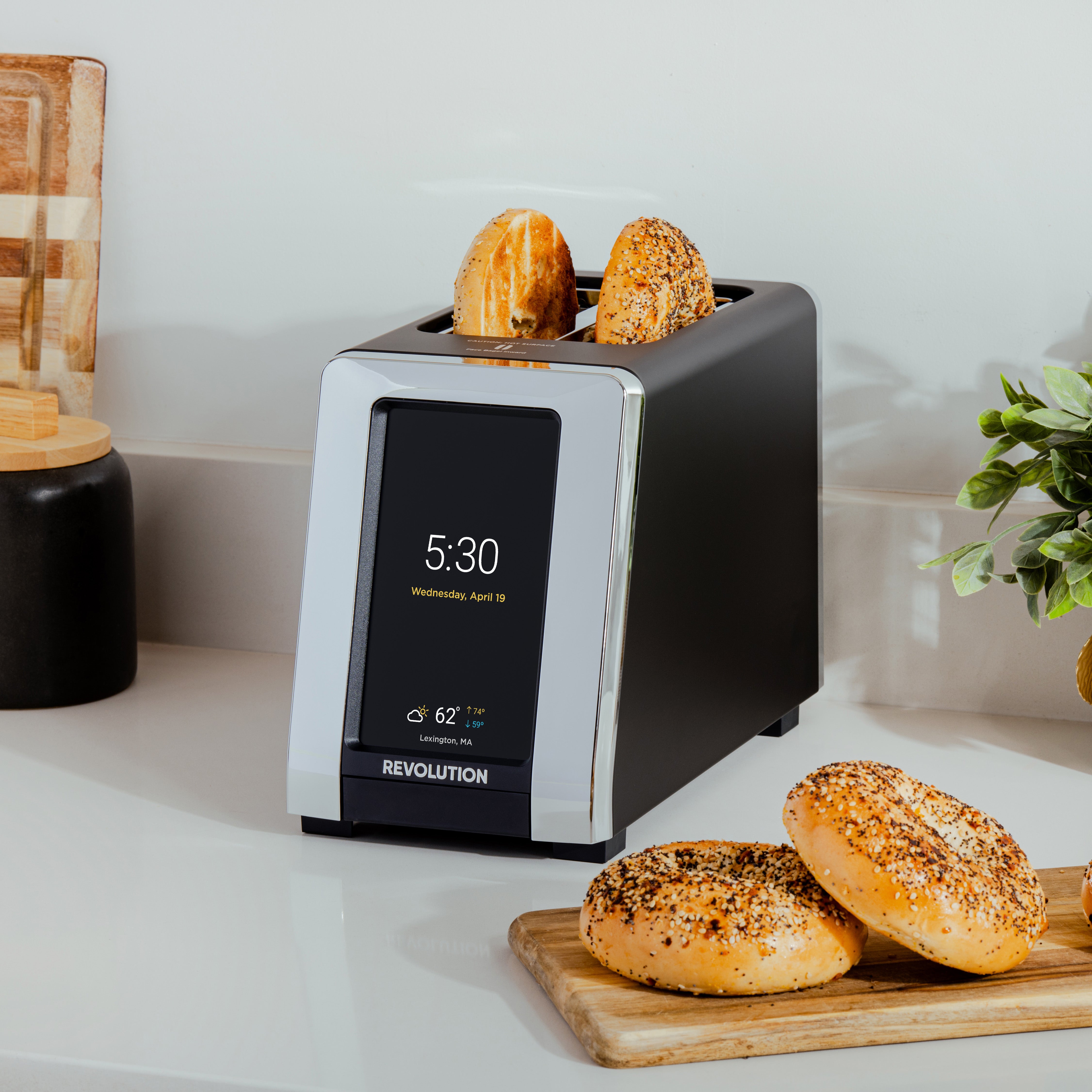 A modern toaster with a digital display shows the time as 5:30, with two pieces of bread being toasted. Nearby, various bagels are placed on a wooden board on a kitchen countertop.