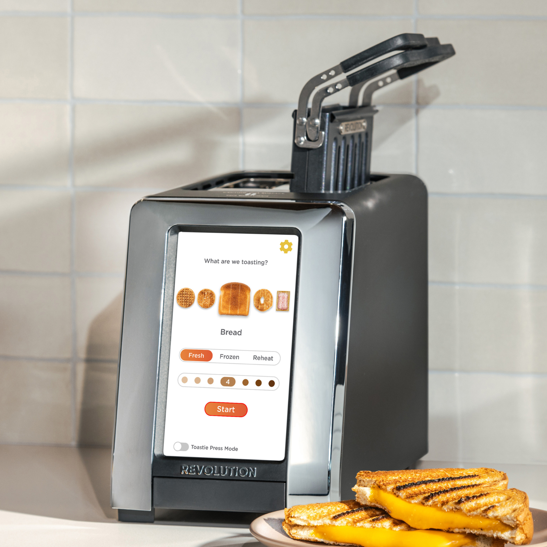 The Revolution Cooking R180 Connect Matte Black + Toastie Press toaster features a touchscreen display with bread options and settings. Its lid is open, and two grilled cheese sandwiches are on a plate in front of a tiled wall.