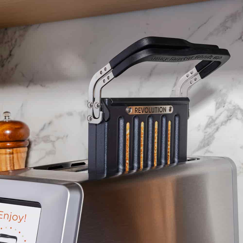 The Revolution Cooking R180 Connect Plus Smart Toaster + Toastie Press, featuring a black "REVOLUTION" handle, is inserted into a sleek stainless steel toaster set before a marble backsplash with a pepper grinder partly visible on the left.