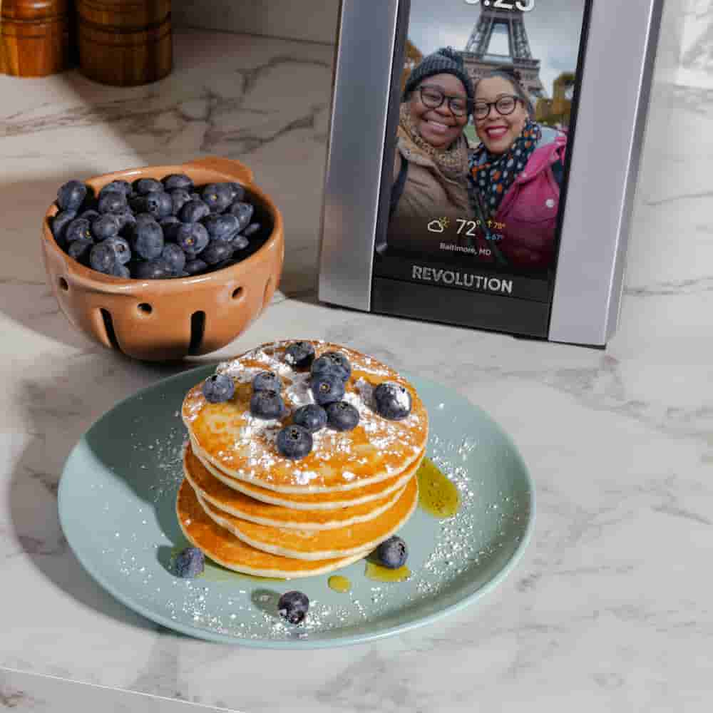 A plate of pancakes with blueberries, syrup, and powdered sugar sits beside a bowl of berries and the Revolution Cooking R180 Connect Plus Smart Toaster + Toastie Press on a marble counter.