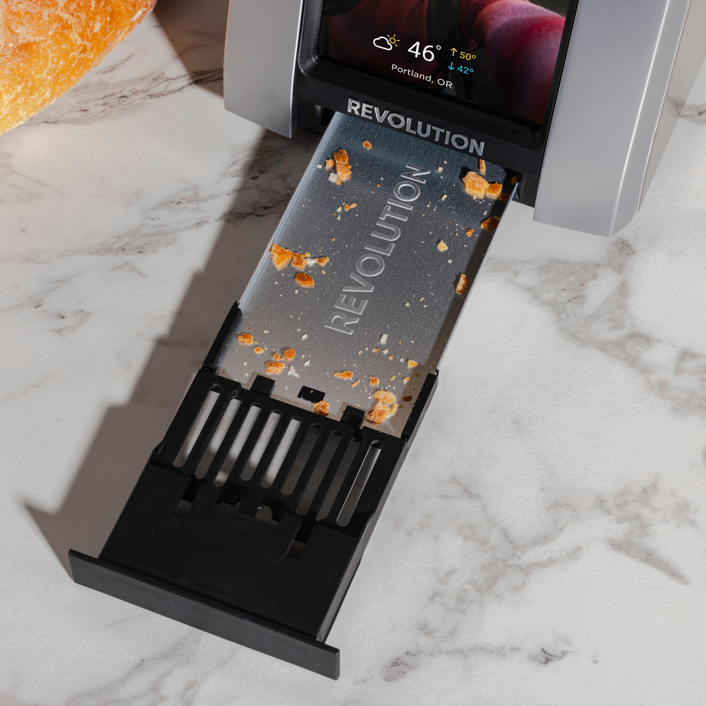 A tray from the Revolution Cooking R180 Connect Plus Smart Toaster is pulled out, revealing bread crumbs. The screen on this Connected Toaster displays Portland's weather. Positioned on a marble countertop, it suggests customized toasting with part of a loaf visible—blending technology with InstaGLO 2.0 efficiency.
