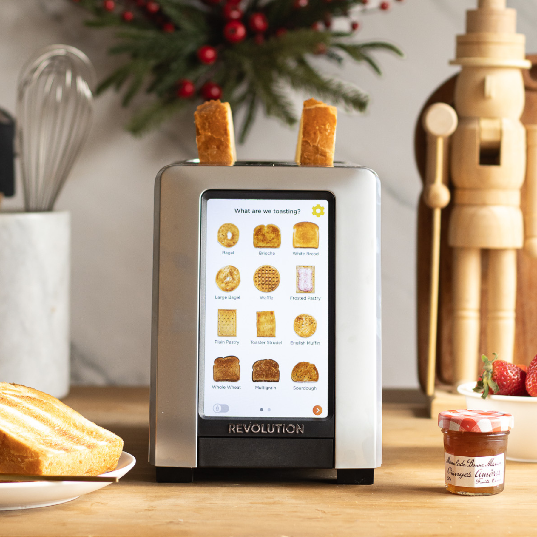 The Revolution Cooking R180 Connect Plus Smart Toaster with Toastie Press and Warming Rack toasts two slices with a touchscreen for bread choices. Sliced bread, jam, strawberries, utensils, holiday decor, and a nutcracker are nearby.
