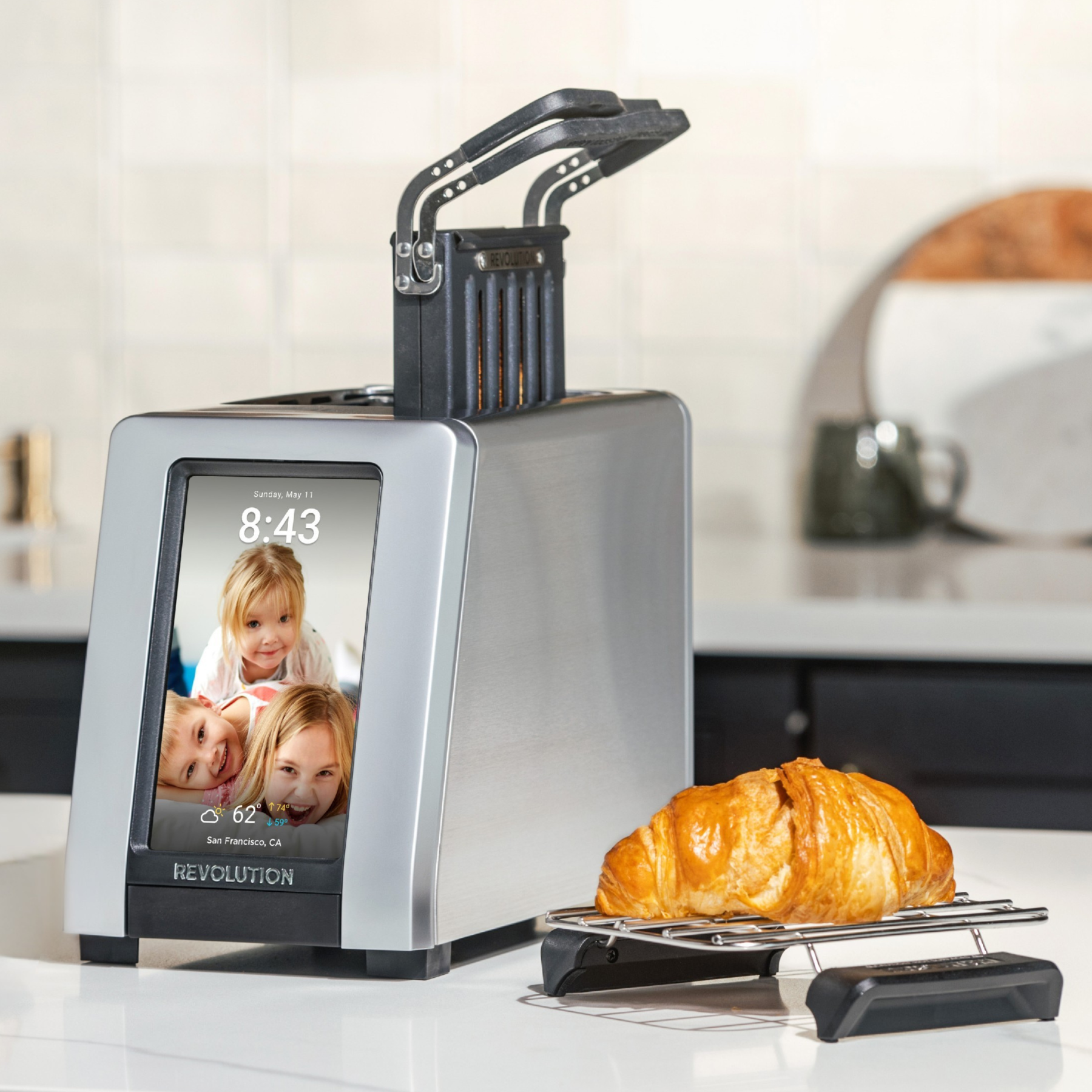 The Revolution Cooking R180 Connect Plus Smart Toaster with digital touchscreen, Toastie Press, and Warming Rack sits on a kitchen counter as a toasted croissant cools nearby.