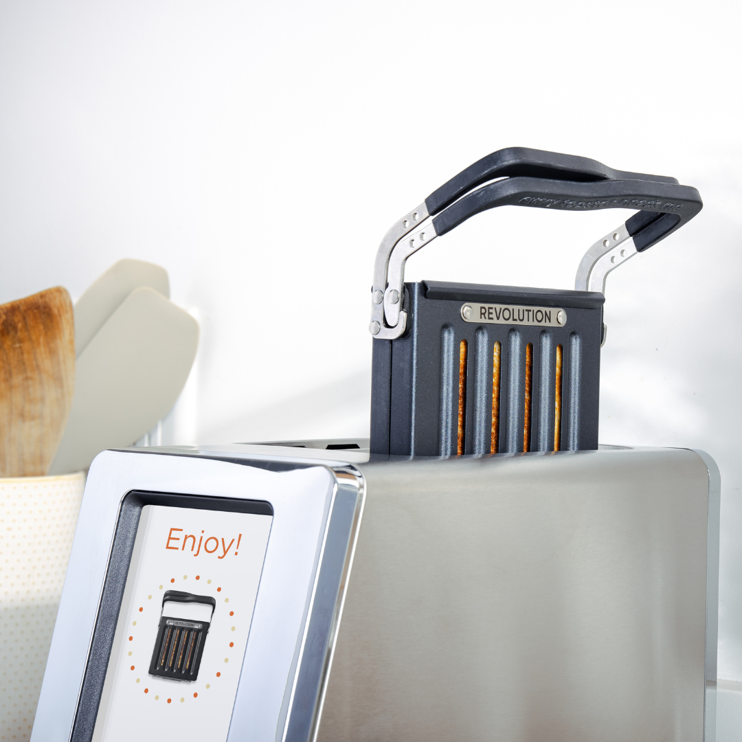 The Revolution Cooking R180 Connect Stainless Steel + Toastie Press, a sleek metallic toaster with a digital “Enjoy!” display and futuristic “Revolution” lever, ejects golden toast as light streams in from the background.