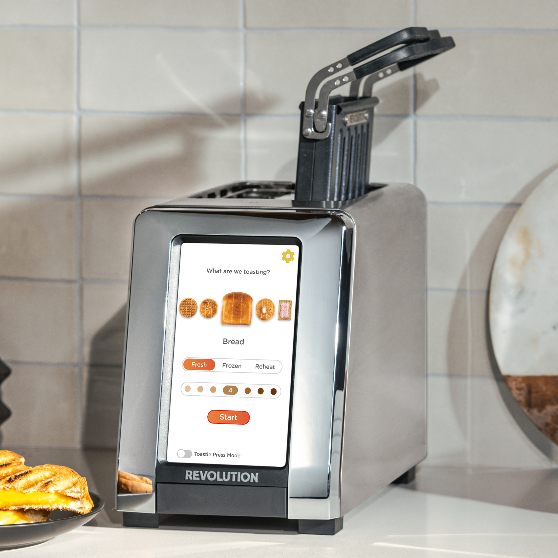 The Revolution Cooking R180 Connect Stainless Steel + Toastie Press features a touchscreen on your kitchen counter, displaying options to toast bread as fresh, frozen, or to reheat.