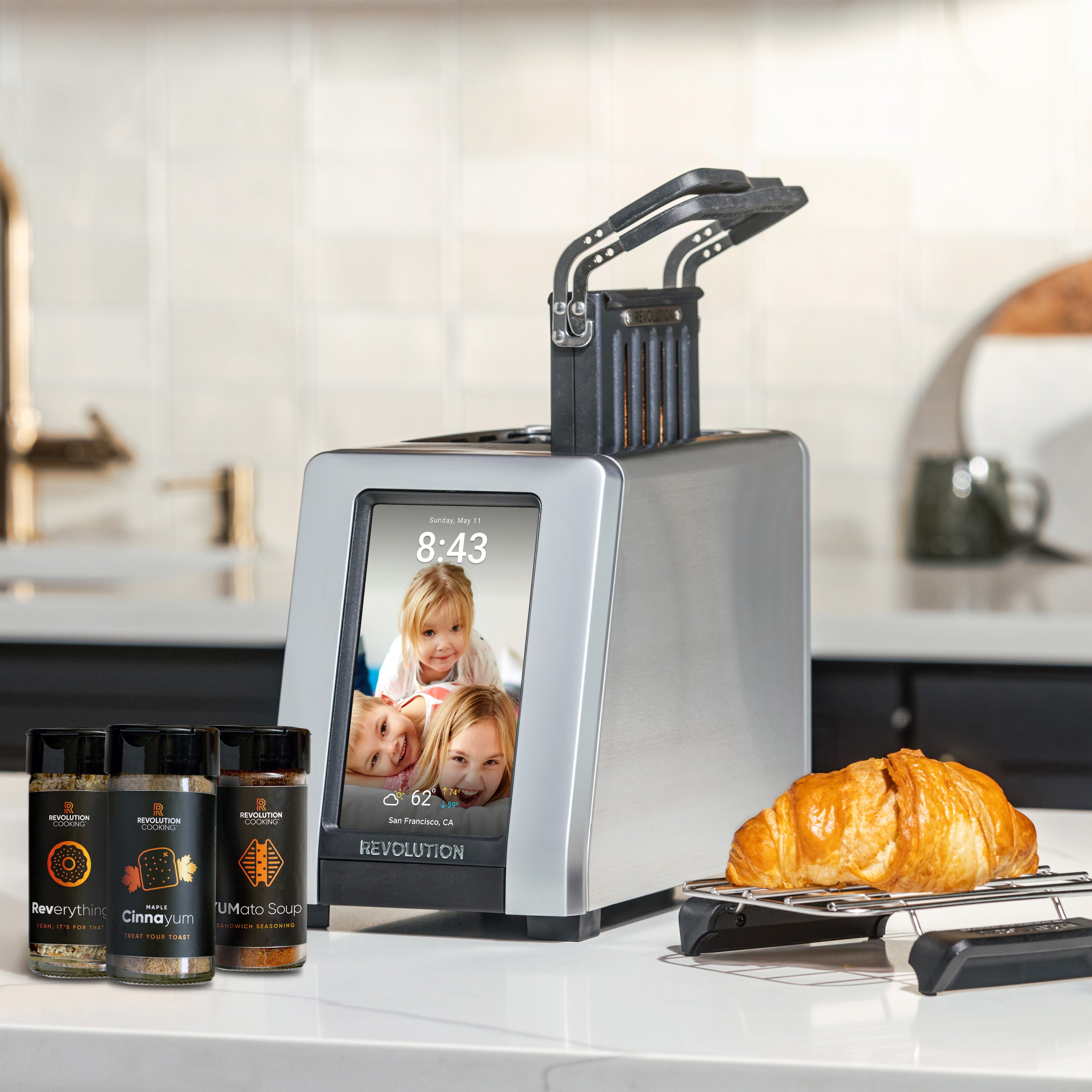 The Revolution Cooking R180 Connect Plus Smart Toaster, Toastie Press, Warming Rack, and 3-Spice Pack sits on the kitchen counter, its touchscreen displaying a family photo. The neutral background highlights the toaster's sleek design.