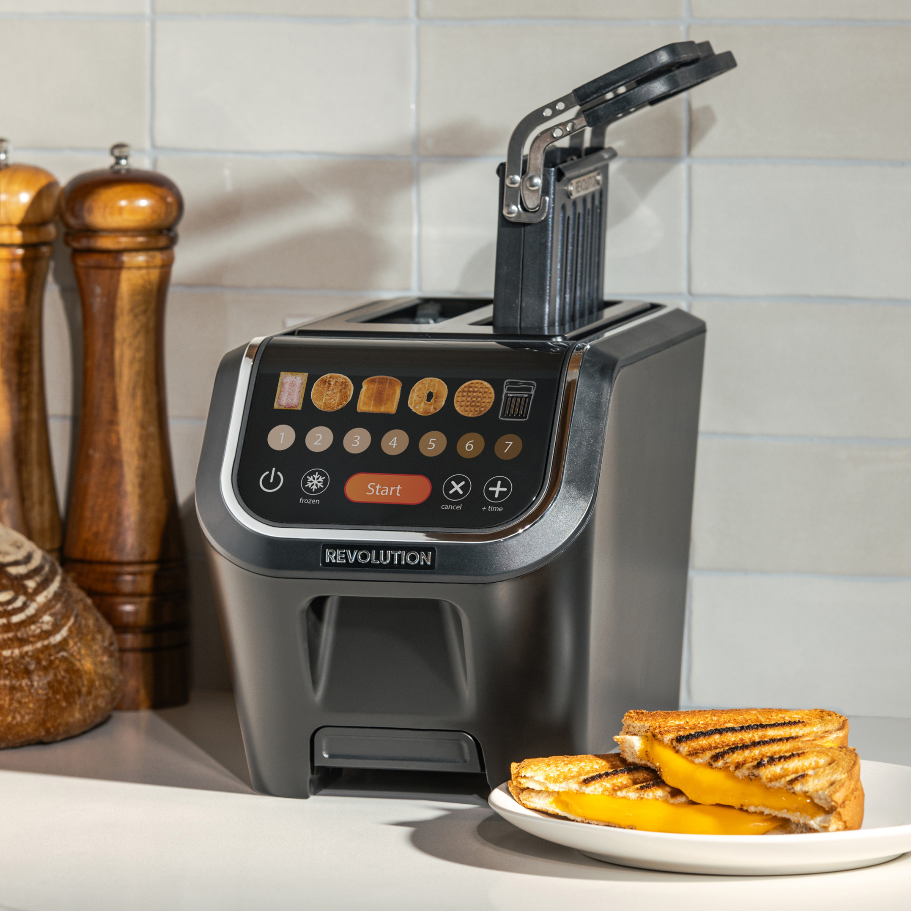 A Revolution Cooking R90 Digital Toaster + Toastie Press with InstaGLO technology sits on the counter beside sliced bread and grilled cheese sandwiches, its slot open. Two pepper grinders are visible in the background.