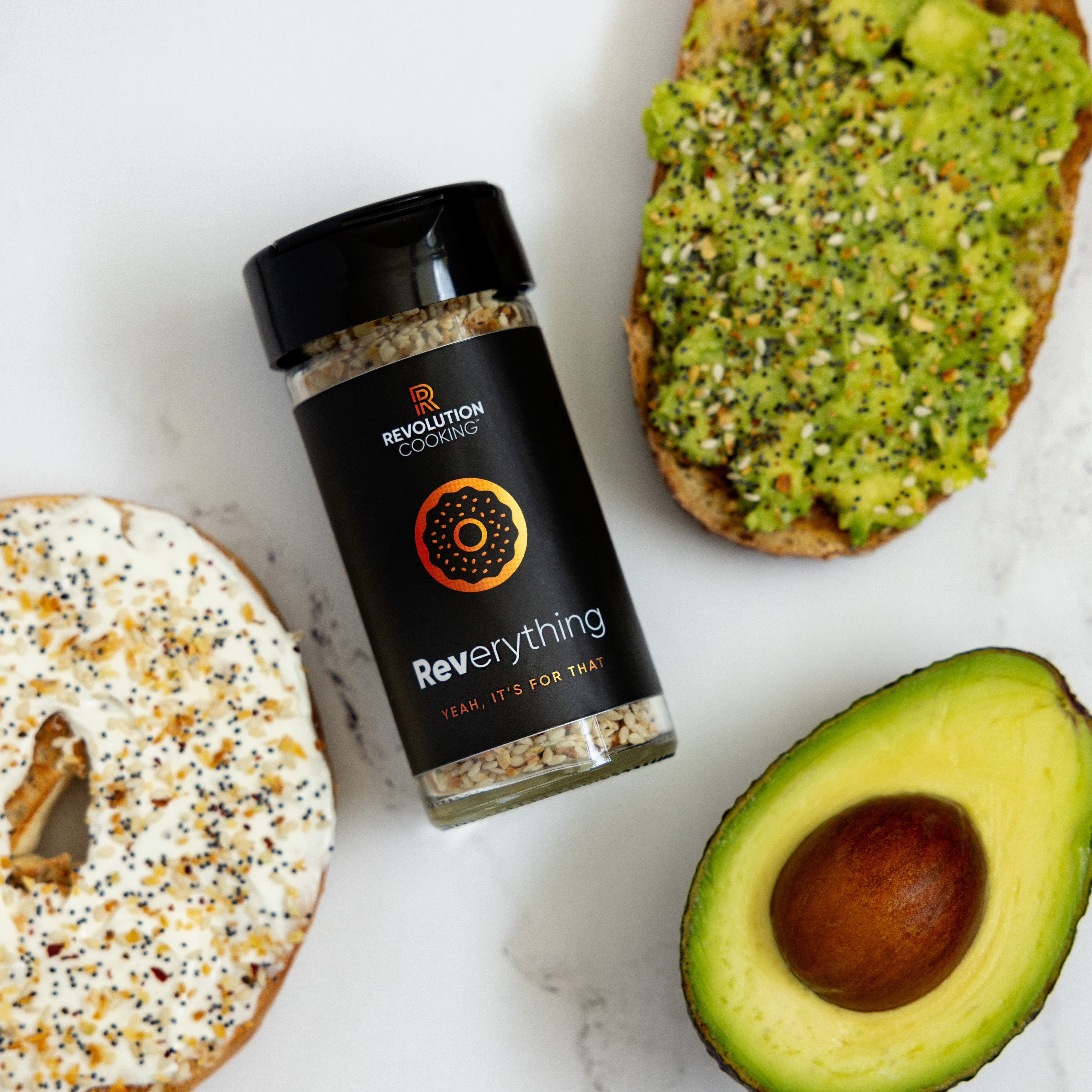 A seasoning jar labeled "REVerything Seasoning" from Ocean State Pepper Co. sits on a marble surface, accompanied by a bagel with cream cheese and seeds, avocado toast seasoned with sumac, and a sliced avocado.