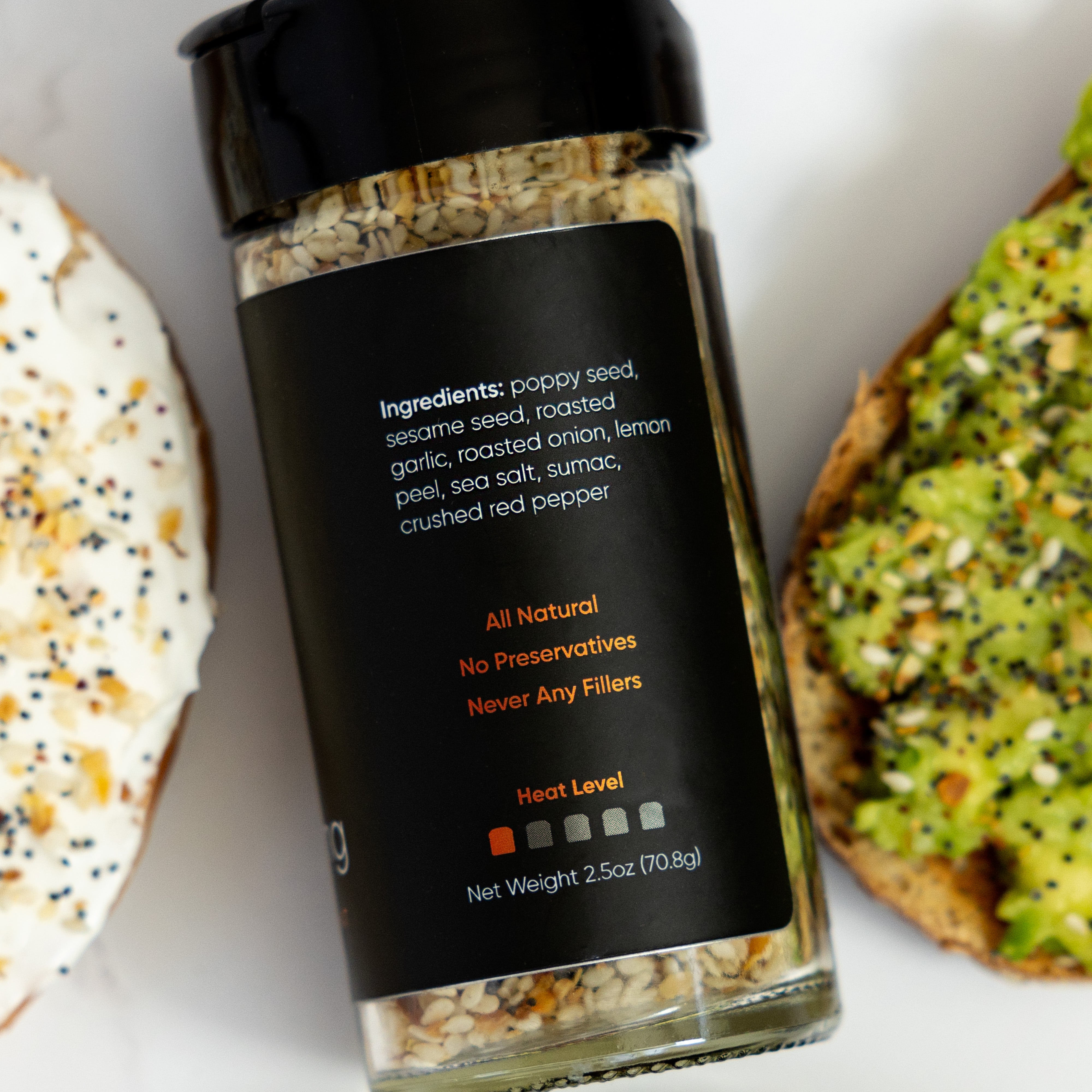 A jar of REVerything Seasoning from Ocean State Pepper Co., which includes ingredients such as poppy seed, sesame seed, and red pepper, is placed beside two slices of bread layered with avocado and cream cheese. The label claims that the product is all-natural and free from preservatives.