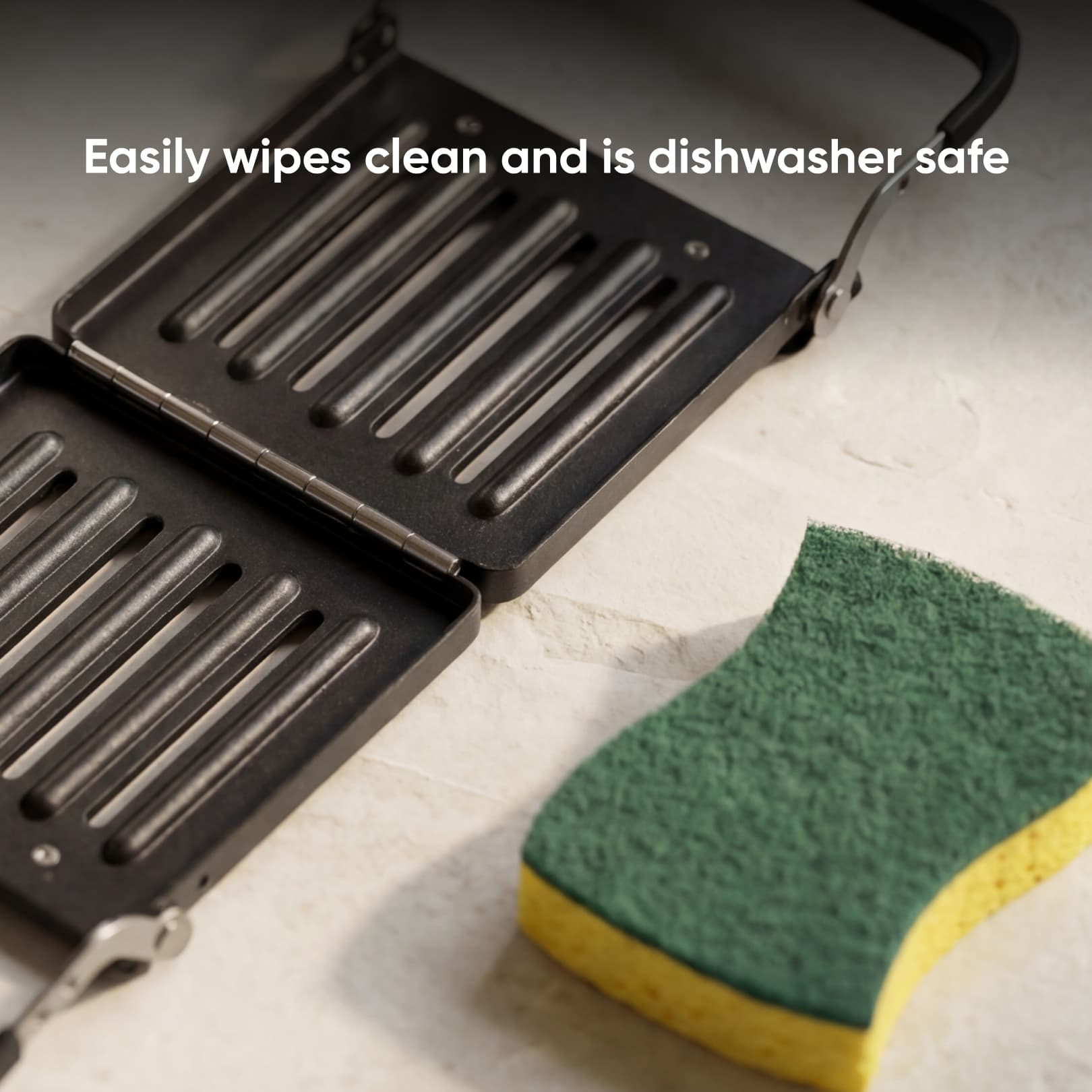 A black Revolution Toastie Press by Revolution Cooking sits open on a countertop next to a yellow and green kitchen sponge. Text above reads, "Easily wipes clean and is dishwasher safe.
