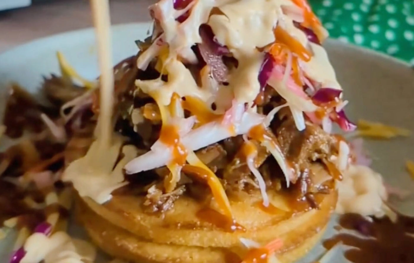 Pulled Pork Sundae stacked on toasted corn cakes with melted cheese, coleslaw, pickled onions, and BBQ sauce.