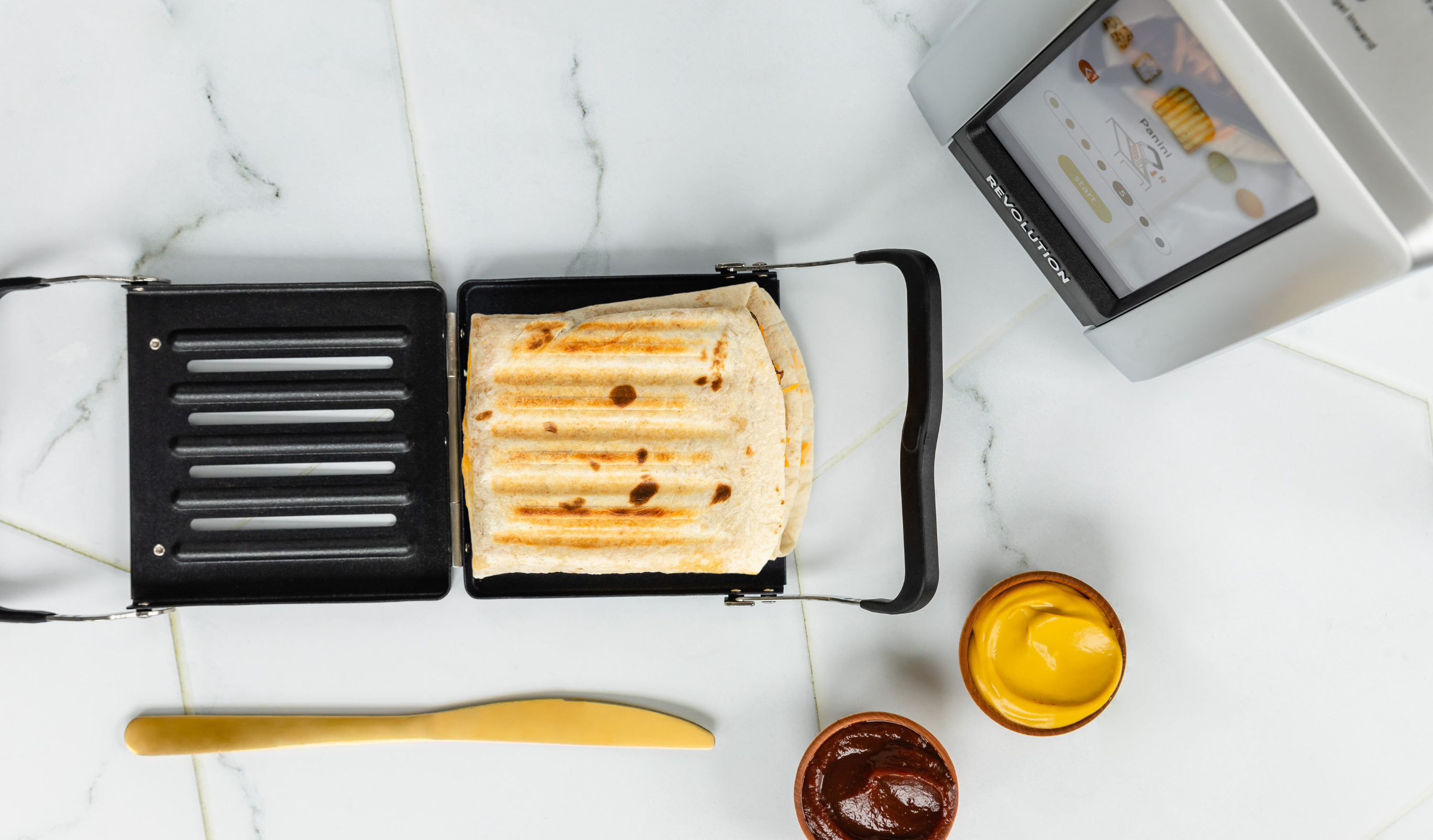 Revolution Toastie Press open on the counter with a toasted pocket fold quesadilla inside. a knife, bbq sauce, and mustard next to it, the toaster is above it