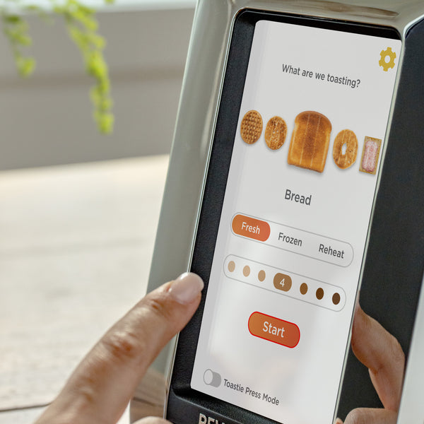 A person is adjusting the toasting options on the Revolution Cooking R180 Connect Smart Toaster's digital screen, displaying bread types and settings like "Fresh," "Frozen," and "Reheat." The InstaGLO® Heating Technology sets the doneness slider to 4, and a "Start" button is located at the bottom.