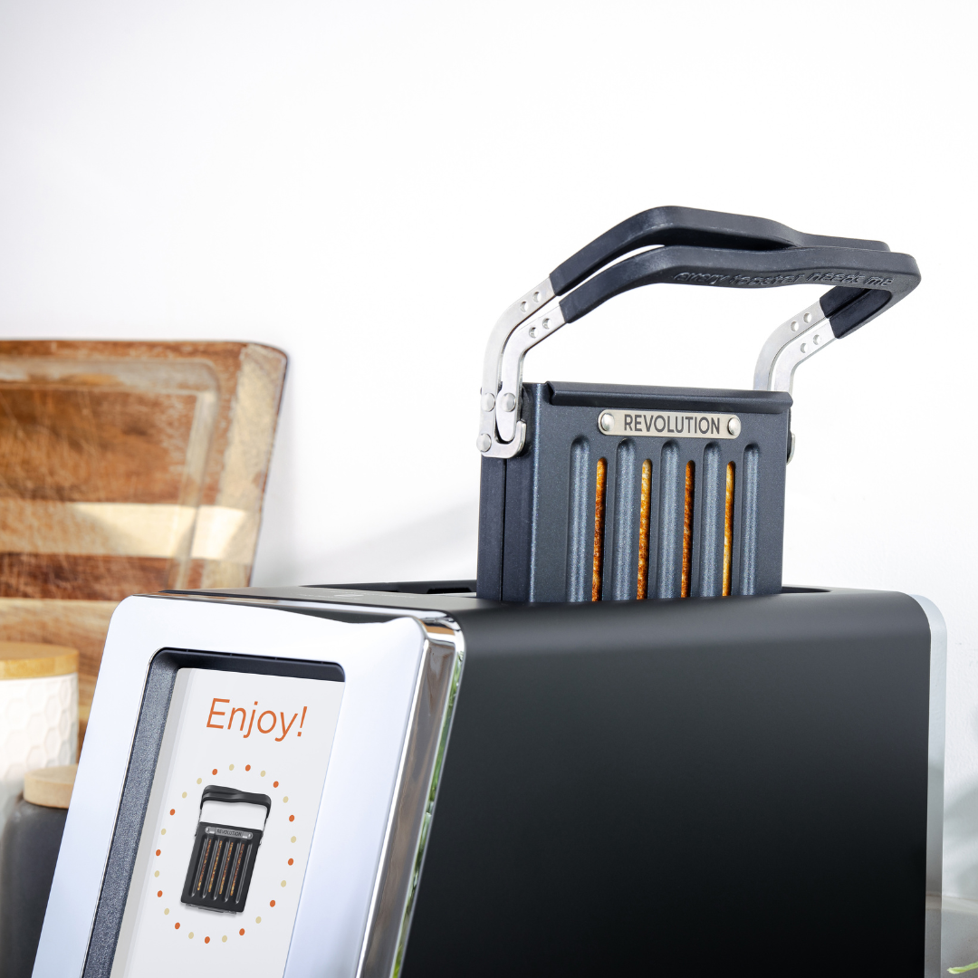 The Revolution Cooking R180 Connect Matte Black + Toastie Press toaster features a digital screen with "Enjoy!" and a "Revolution" lever, shown toasting bread. Cutting boards are in the background.