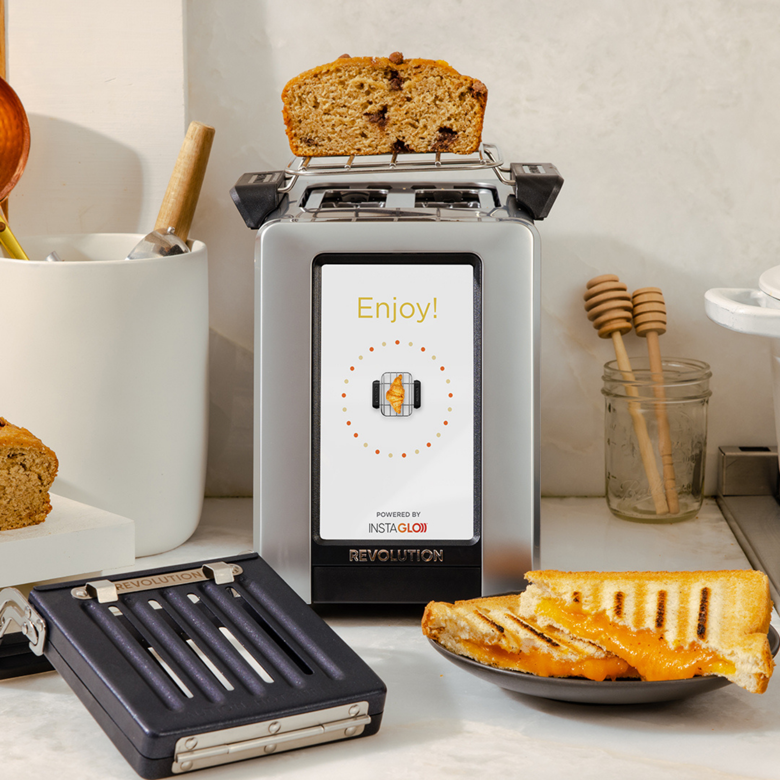 The Revolution Cooking R180 Connect Plus Smart Toaster with Toastie Press and Warming Rack sits on a kitchen counter, toasting bread. Around it are toasted sandwiches, honey jars, and utensils. The digital display reads "Enjoy!.