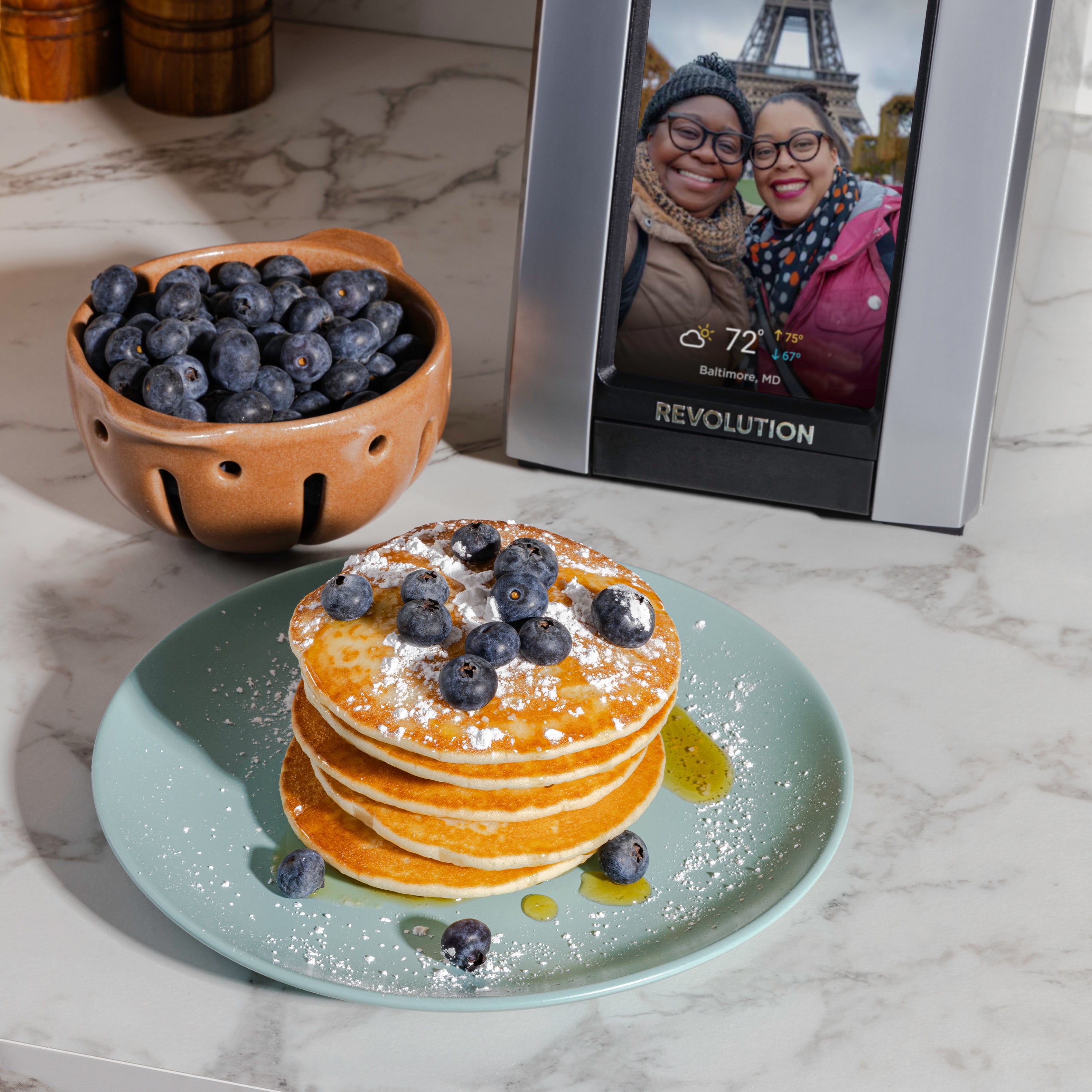 A stack of pancakes crowned with blueberries and powdered sugar rests on a marble countertop, creating a perfect backdrop for the Revolution Cooking R180 Connect Plus Smart Toaster's display in the background, where a digital picture frame shows two people smiling. A bowl of fresh blueberries is positioned next to this breakfast treat.