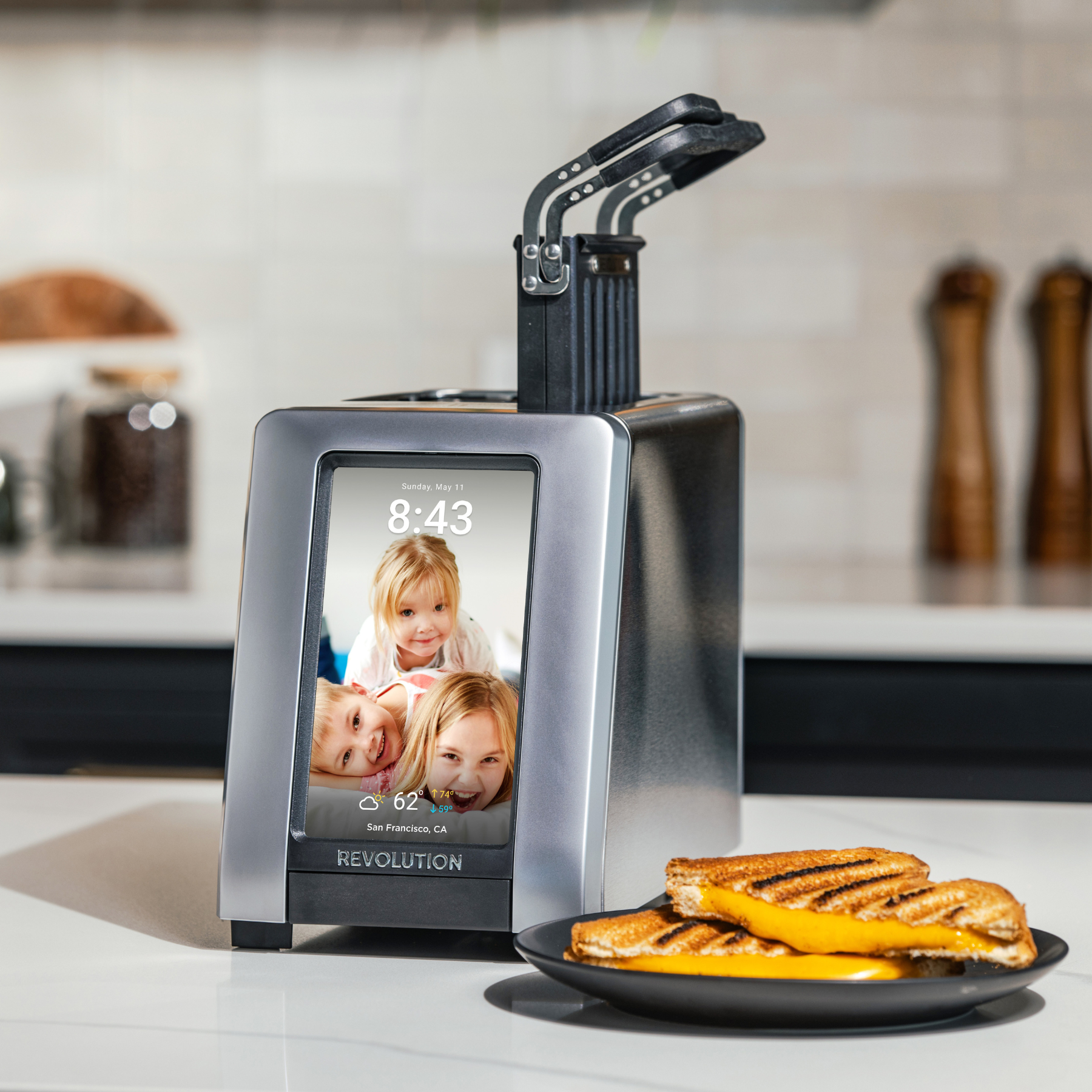 The Revolution Cooking R180 Connect Plus Smart Toaster + Toastie Press, featuring a touchscreen display for family photos, time, date, weather, and location, sits on a kitchen counter beside a plate of toasted, buttered bread slices.
