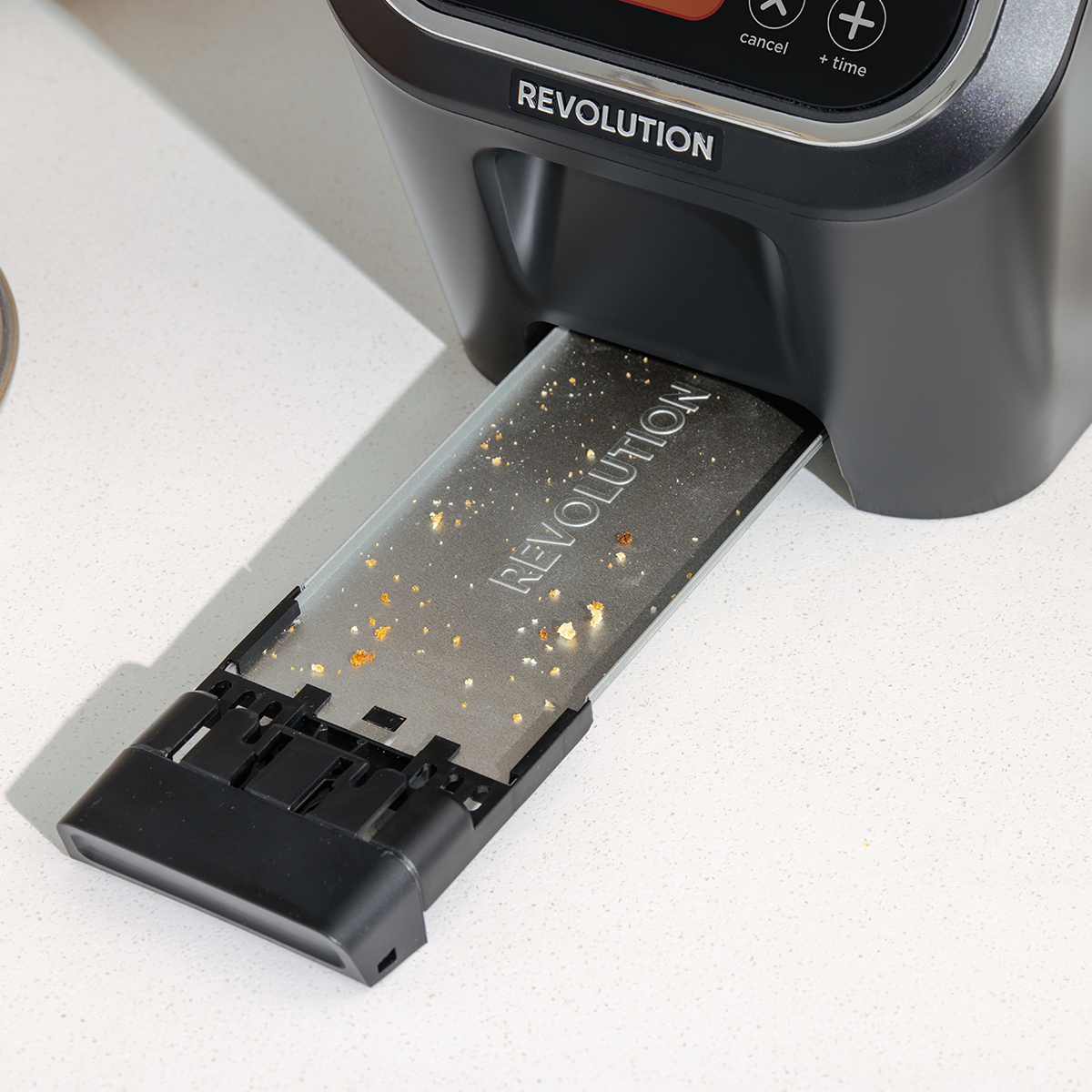 A modern toaster with the brand name "Revolution" is displayed. The crumb tray is pulled out, revealing some crumbs. The design features a sleek, dark finish with a digital interface.