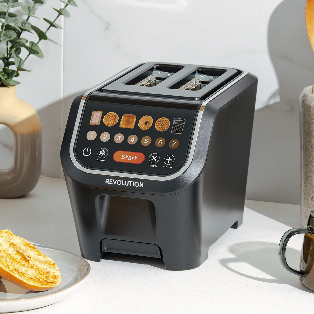 A sleek, modern toaster with touch controls displaying various bread options sits on a kitchen counter. A toasted bagel is on a plate nearby. The toaster has a black finish and "Revolution" branding. A plant and cup are in the background.