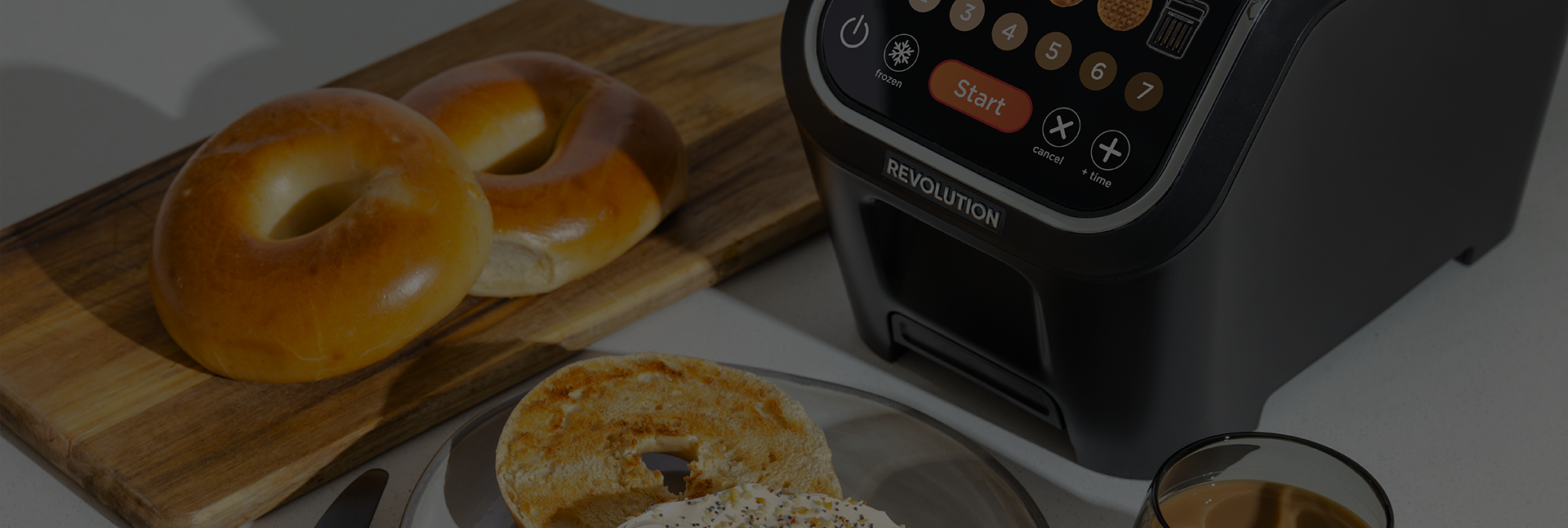 A toaster with illuminated buttons sits on a kitchen counter. Nearby is a wooden board with two plain bagels and a plate with a toasted bagel half topped with cream cheese. A small glass of coffee is also visible.