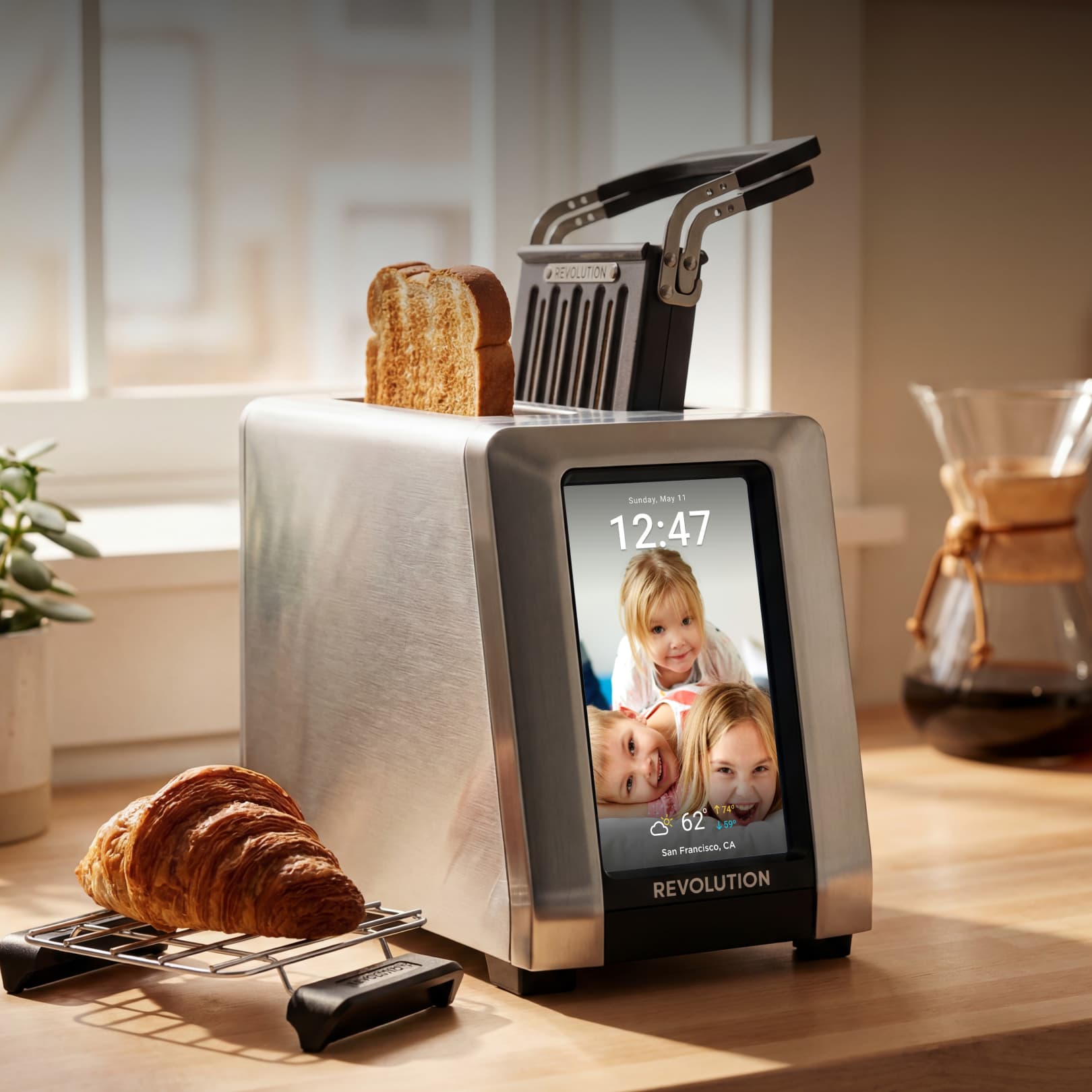 The Revolution Cooking R180 Connect Plus Smart Toaster with Toastie Press and Warming Rack features a digital touchscreen showing family photos and the time. Slices of bread are inside, croissants cool on the rack, and coffee sits in the background.
