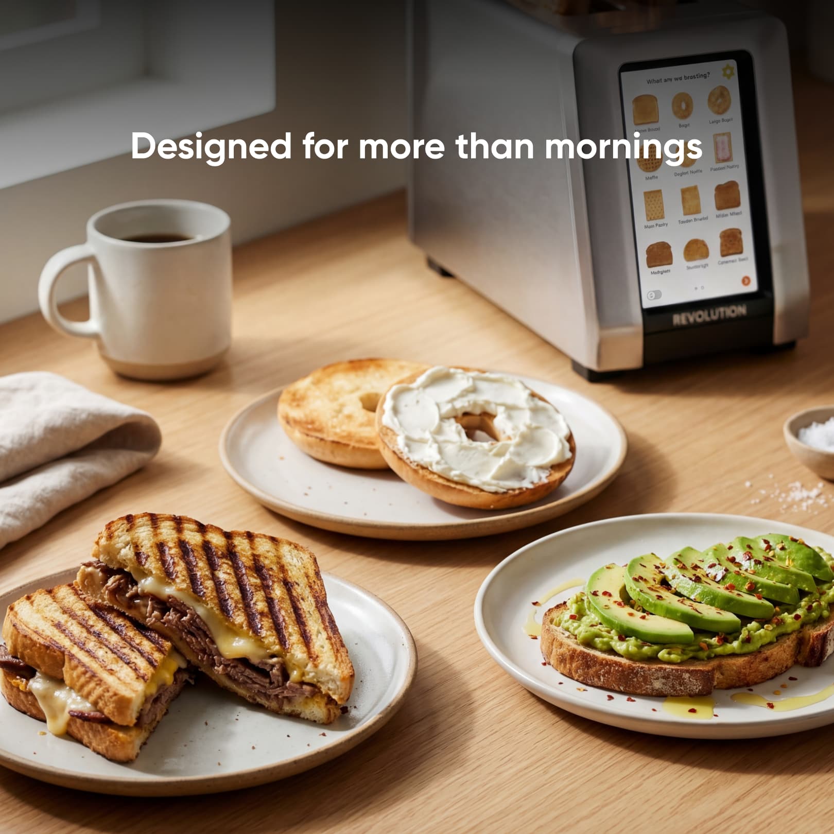 Three plates with breakfast foods—grilled cheese, avocado toast with red pepper flakes, and a bagel with cream cheese—sit beside a coffee mug and the Revolution Cooking R180 Connect Plus Smart Toaster + Toastie Press + Warming Rack.