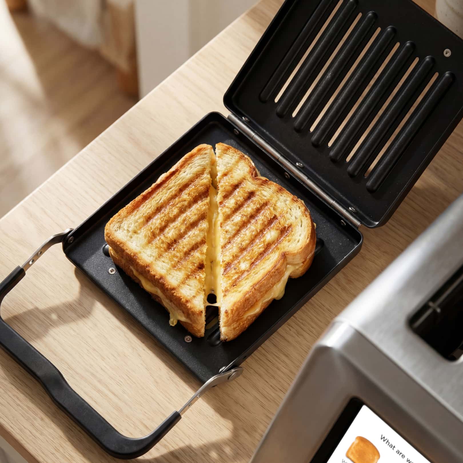 A golden-brown grilled cheese sandwich, cut diagonally with melted cheese at the edges, sits inside the open Revolution Cooking Revolution Toastie Press on a light wooden countertop.
