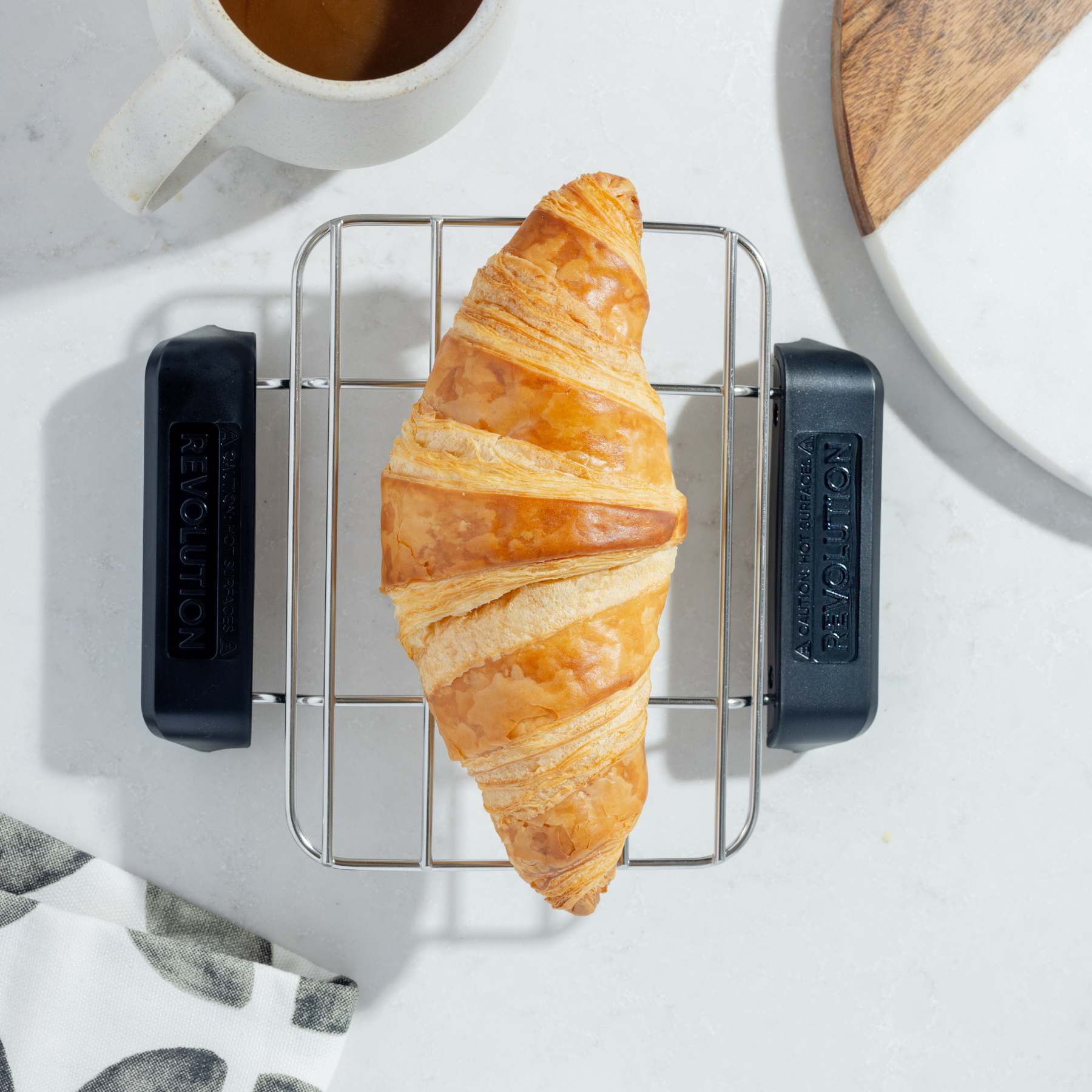 A golden croissant rests on the Revolution Cooking R180 Connect Plus Smart Toaster's warming rack, next to coffee and a cutting board on a white countertop. A black and white napkin is partially visible.